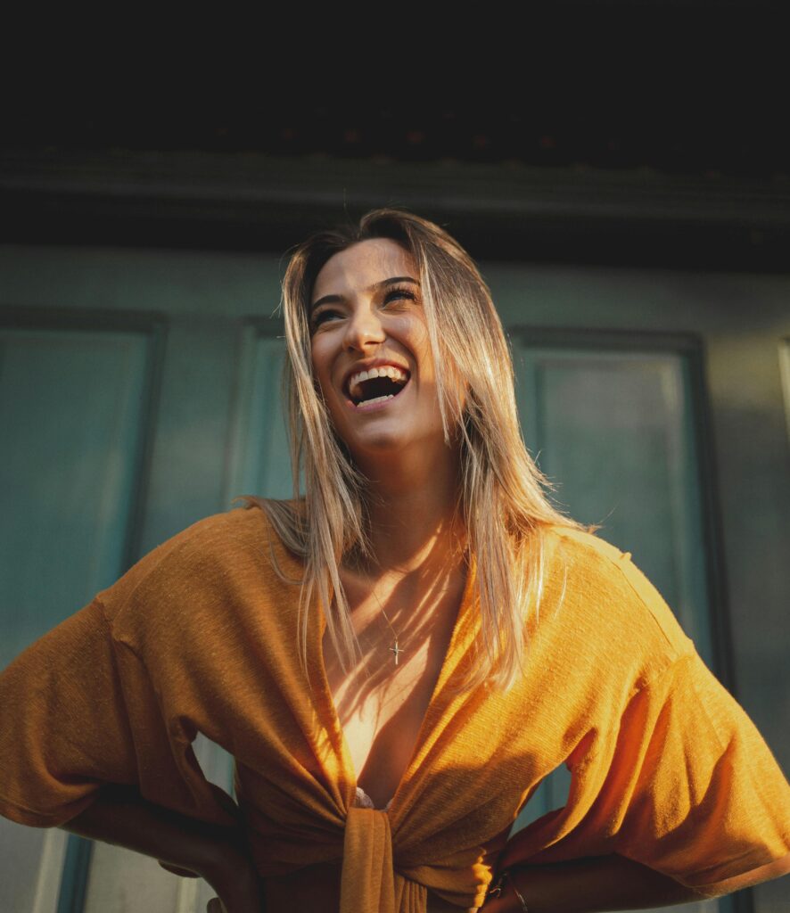 A cheerful woman with blonde hair laughing and posing outdoors in a stylish yellow blouse.