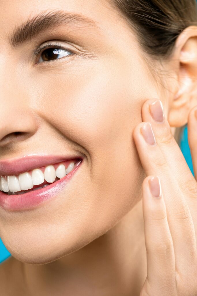A close-up portrait showing a woman gently touching her cheek, showcasing healthy skin and a vibrant smile.