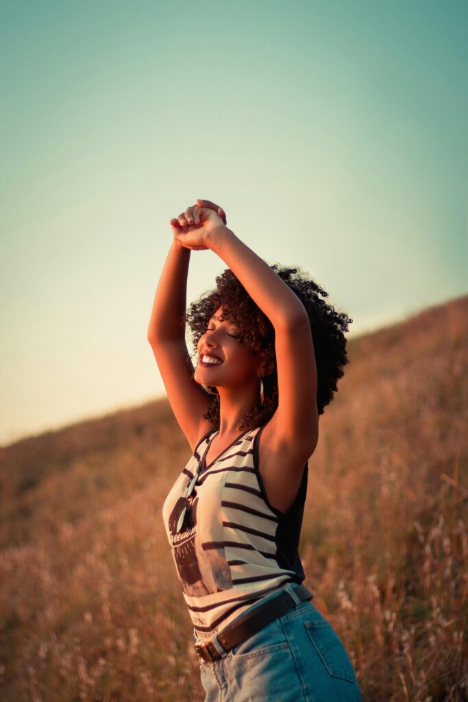 Smiling young African American female in jeans and striped tank top with closed eyes and raised hands enjoying nature