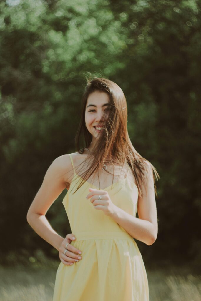 Young brunette woman smiling while wearing a yellow dress in a sunny outdoor setting.