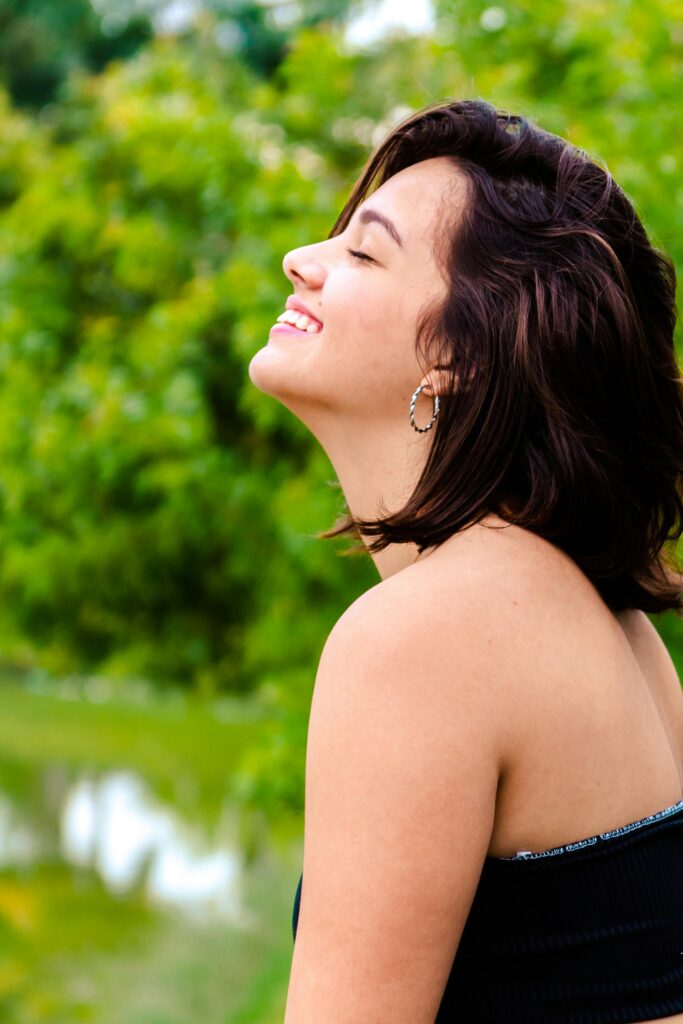 A joyful woman with short brunette hair smiles outdoors in a park, surrounded by lush greenery.